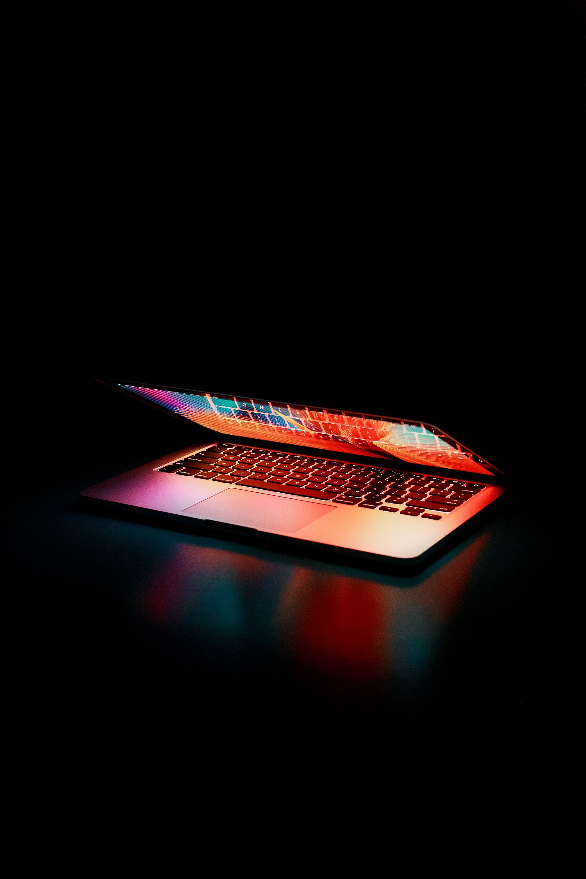 A laptop in darkness, its keyboard glowing with vibrant, multicolored lights, casting a soft, colorful reflection on the surface.