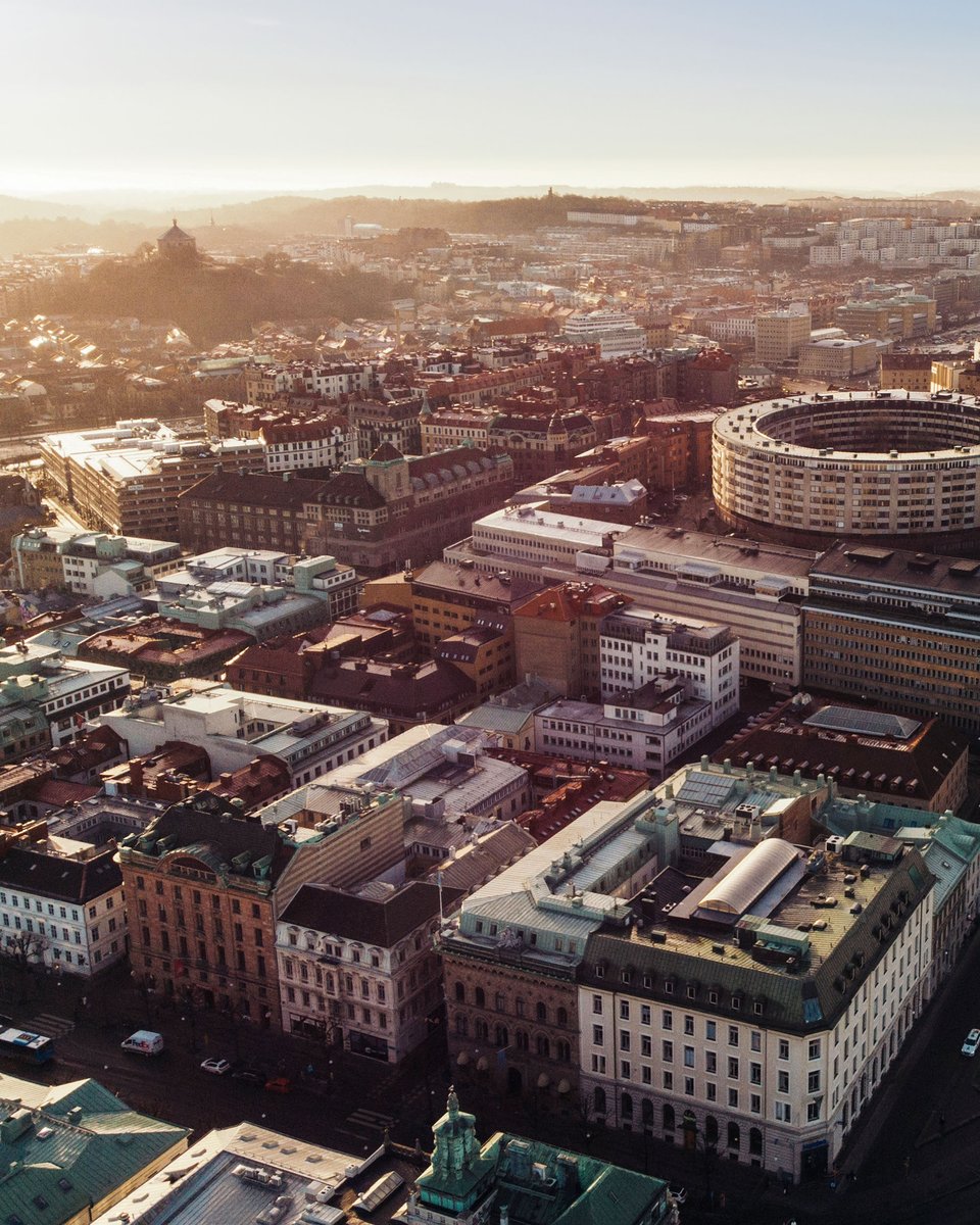 Aerial view of a city with diverse architecture, a circular building, river, bridges, under the warm glow of the setting or rising sun.